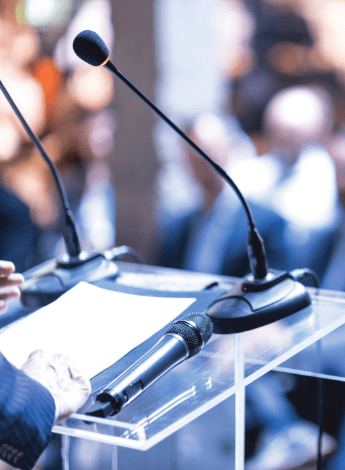 The image is of a podium. There are two microphones attached to it and another handheld microphone on top, along with a piece of paper. Someone is standing at the podium but is out of frame.